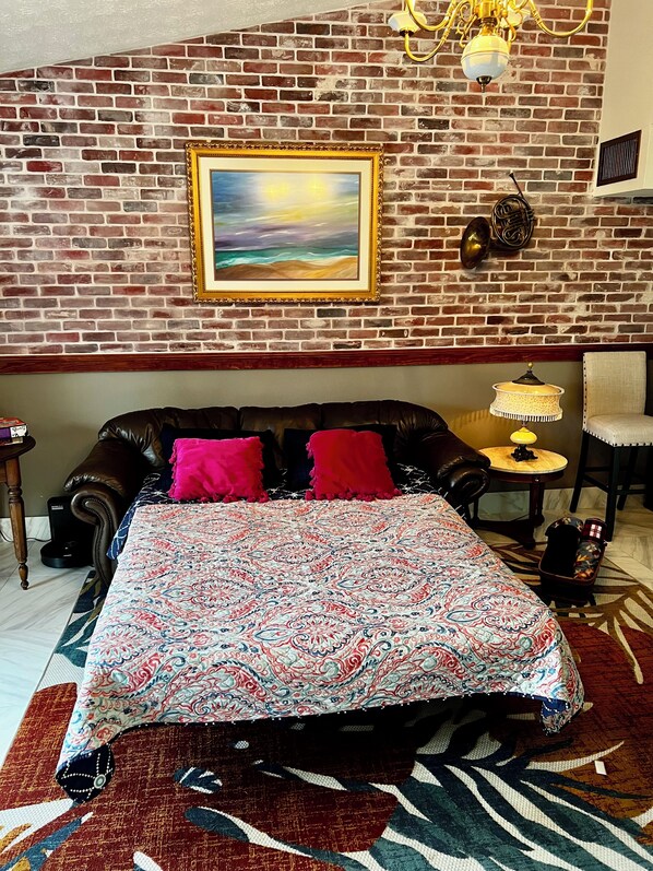 Quarto com parede de tijolos, cama com colcha estampada, mesa pequena com abajur e uma cadeira.