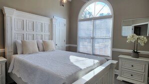A bedroom with a large bed, a nightstand with a lamp, a dresser, a mirror, and a window with blinds.