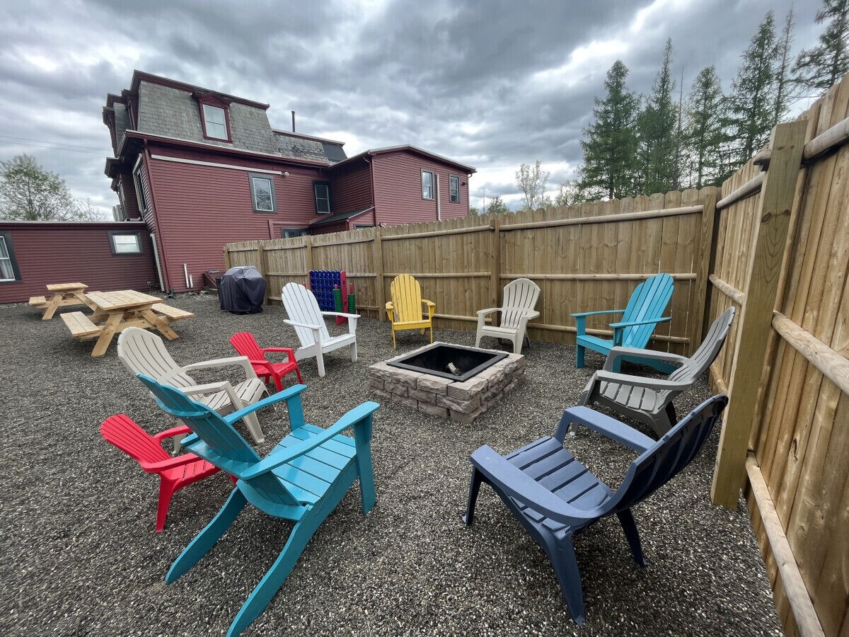 A backyard with a fire pit surrounded by colorful Adirondack chairs, a wooden fence, and a red building in the background.