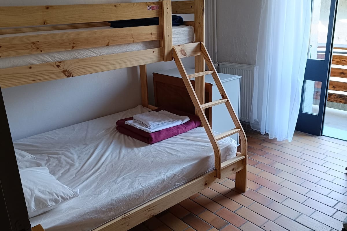 Ein Stockbett mit Holzleiter, ein kleiner Holztisch und ein Fenster mit Vorhängen.