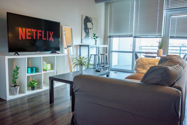 Una sala de estar moderna con un televisor de pantalla plana mostrando 'NETFLIX', un sofá, una mesa de centro y una estantería con plantas.