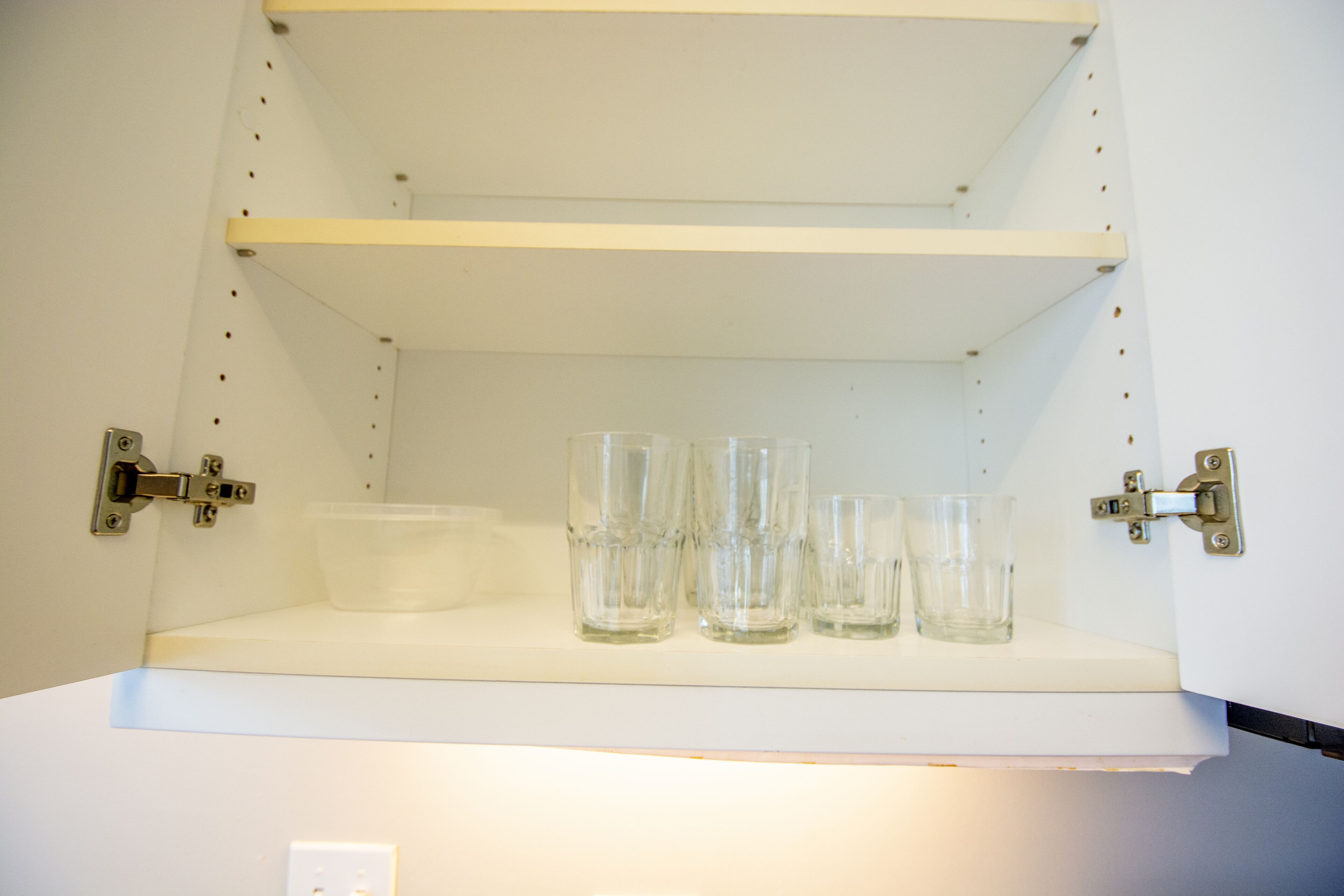 A white shelf with four clear glasses and a clear plastic container.