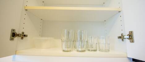 A white shelf with four clear glasses and a clear plastic container.