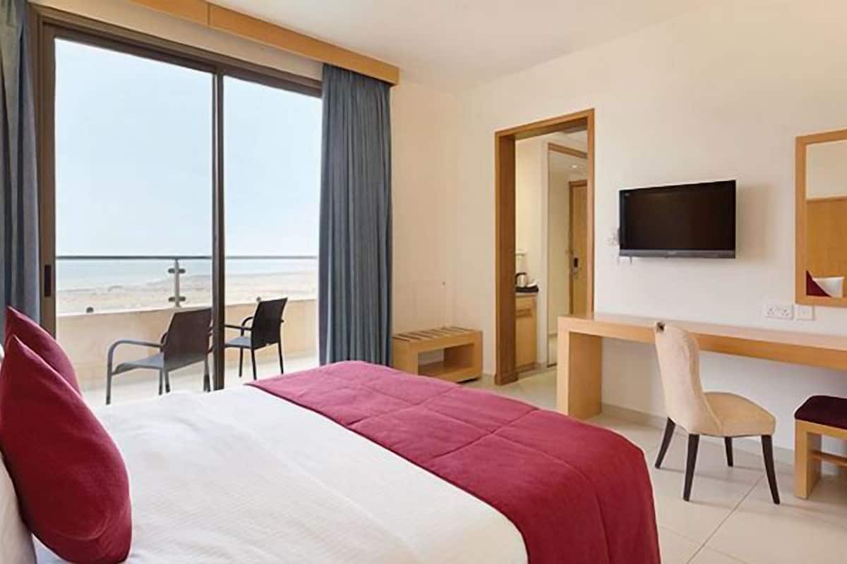 A hotel room with a bed, a desk, a chair, a TV, and a view of the beach.