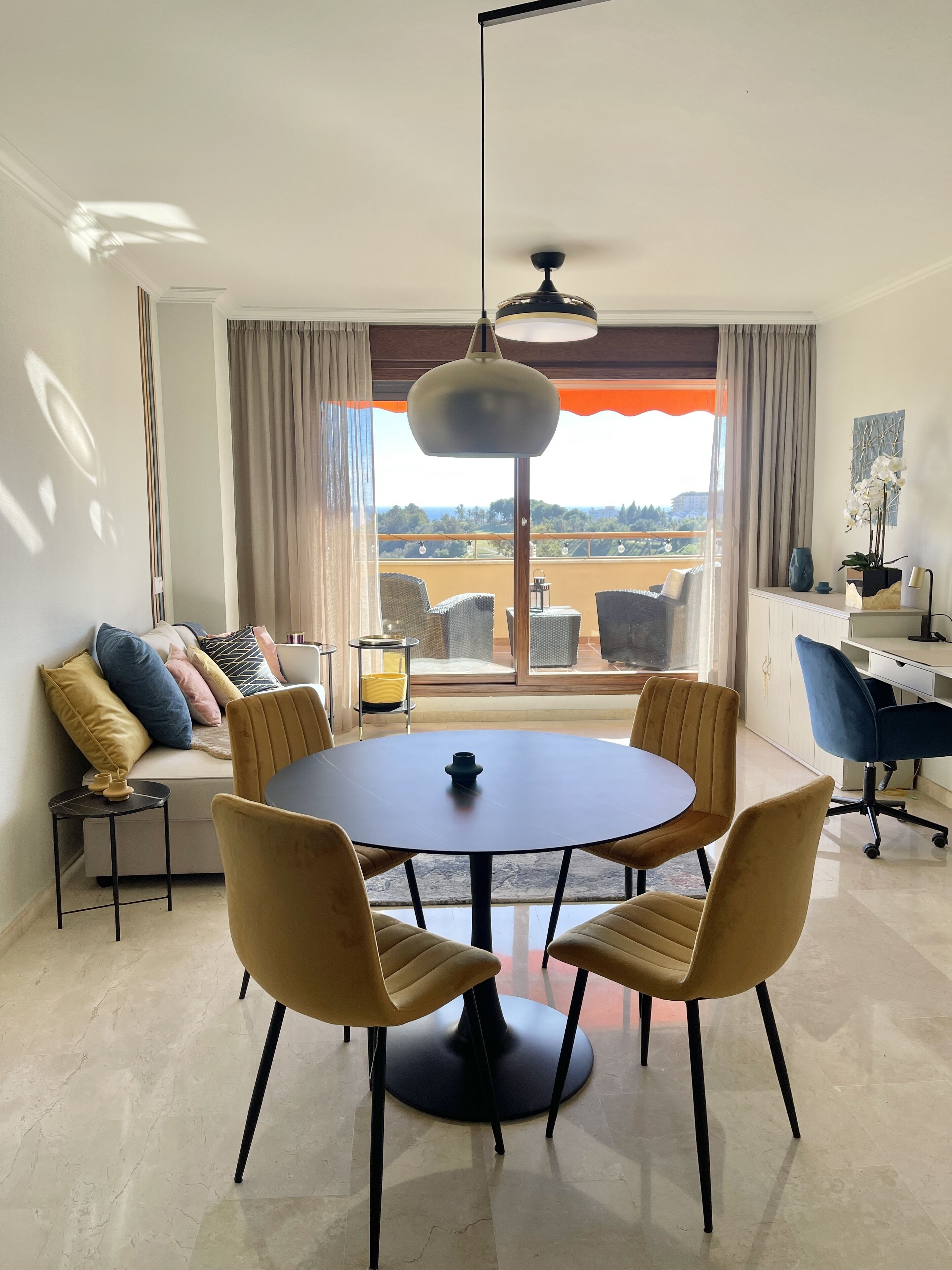 A modern living room with a round table, four chairs, a sofa with cushions, a ceiling fan, and a large window with a view of the outdoors.