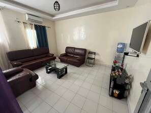 A living room with two brown leather sofas, a glass coffee table, a TV mounted on the wall, and a small side table with a water dispenser and decorative items.