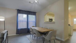 Een eethoek met een houten tafel en stoelen, een groot raam met gordijnen, en een keuken in de achtergrond.