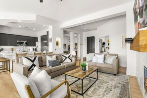 Ampla sala de estar com sofá, poltronas e mesa de centro. Uma cozinha é visível ao fundo.
