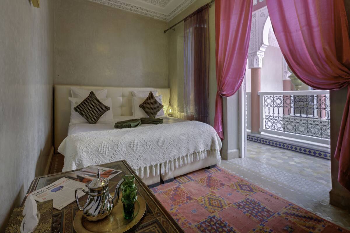 A hotel room with a bed, a table with a teapot and glasses, and a view of the outside balcony.