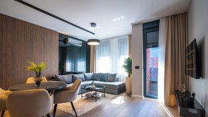 Una sala de estar moderna con un sofá, una mesa de centro y sillas. Hay un televisor montado en la pared y una ventana con cortinas.