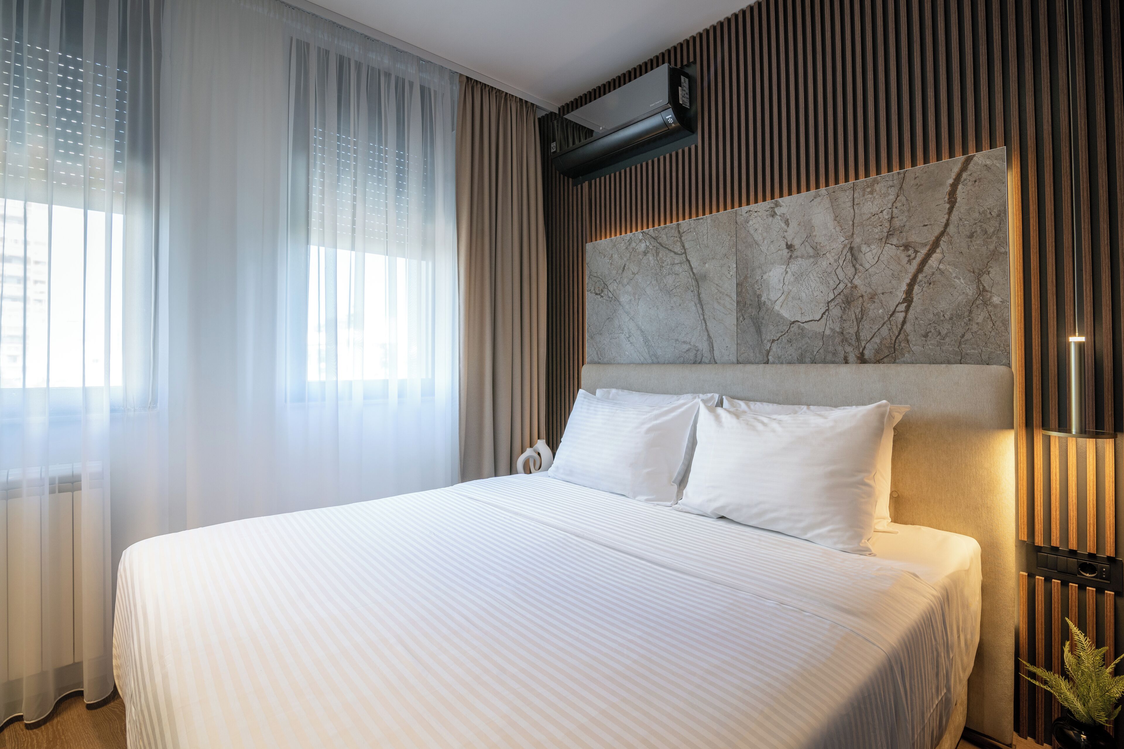 A hotel room with a large bed, a marble headboard, a wall-mounted air conditioner, and a potted plant.