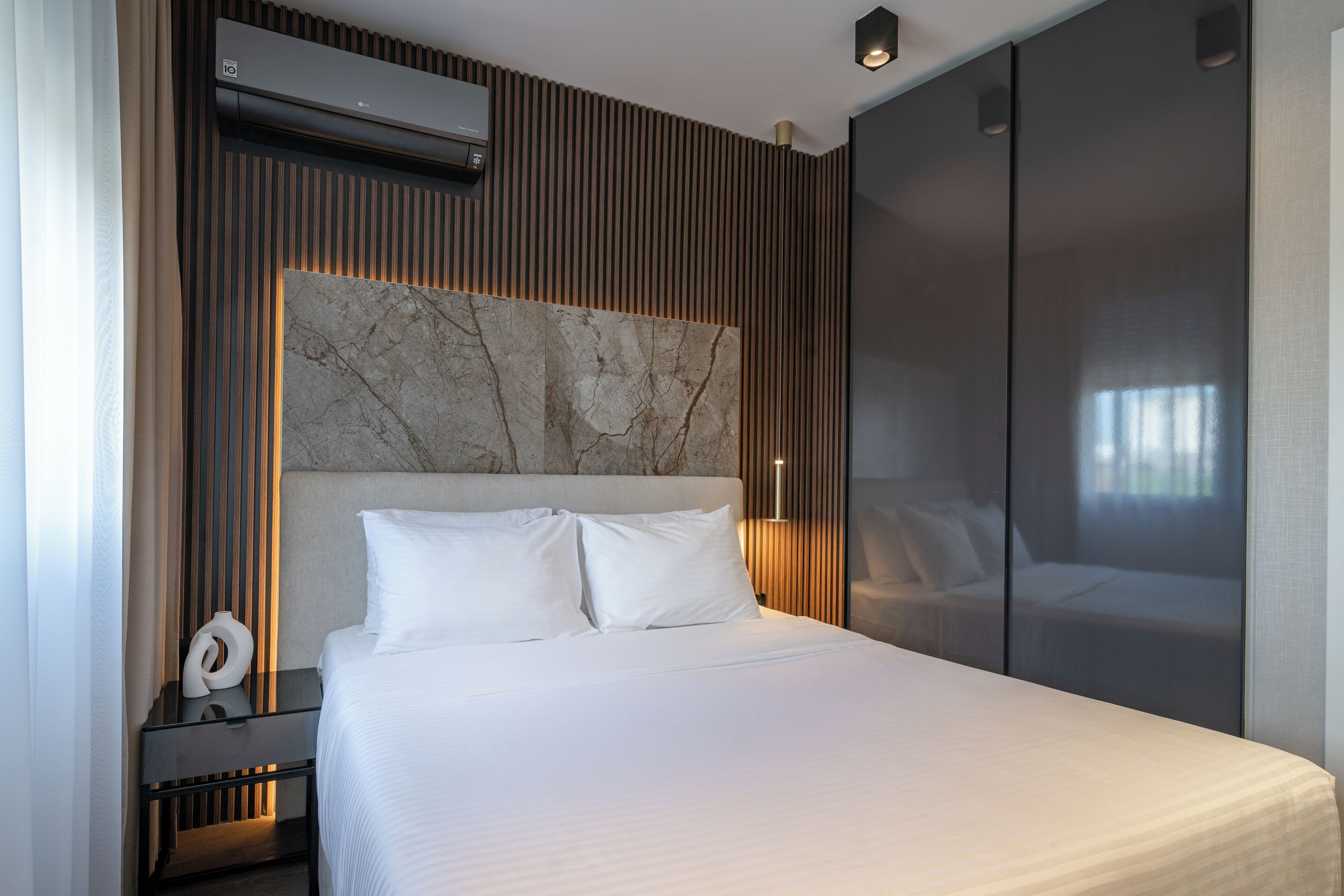 A modern hotel room with a large bed, a marble headboard, a wall-mounted air conditioner, and a nightstand with a lamp.