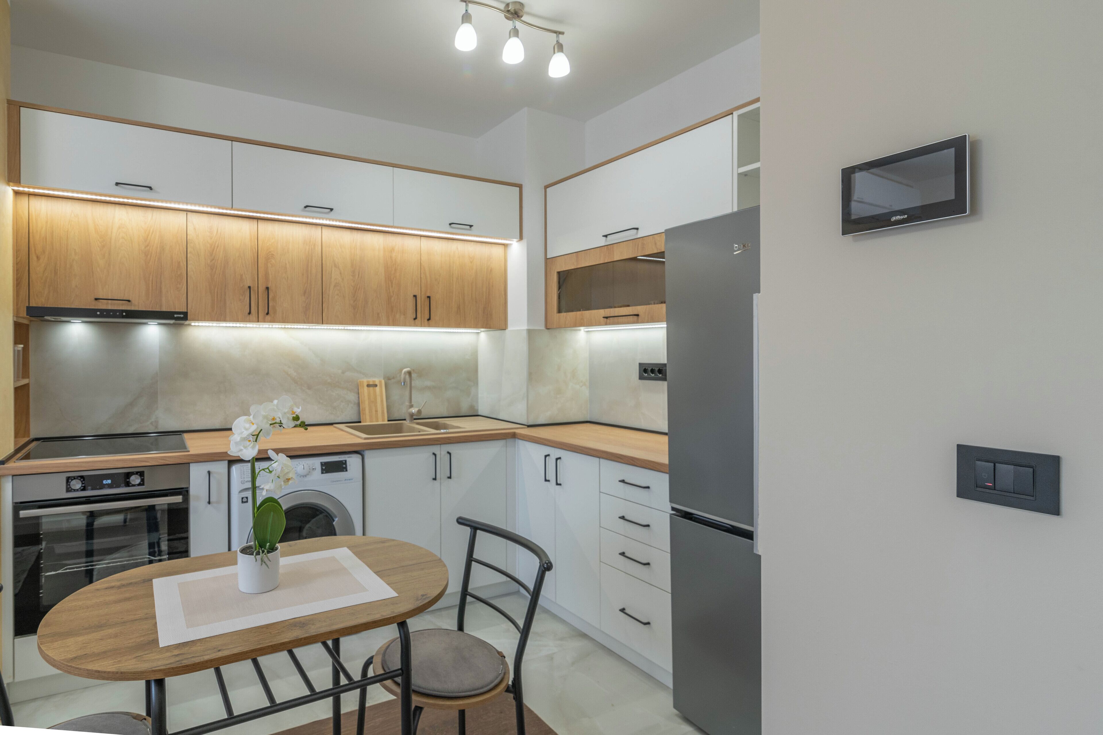 A modern kitchen with wooden cabinets, a stainless steel refrigerator, a built-in oven, and a microwave. There is a small table with a chair and a vase of flowers.