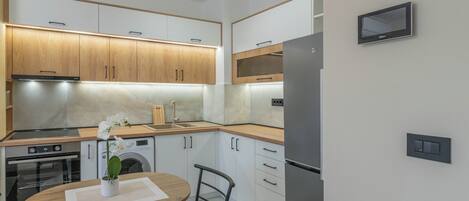 A modern kitchen with wooden cabinets, a stainless steel refrigerator, a built-in oven, and a microwave. There is a small table with a chair and a vase of flowers.