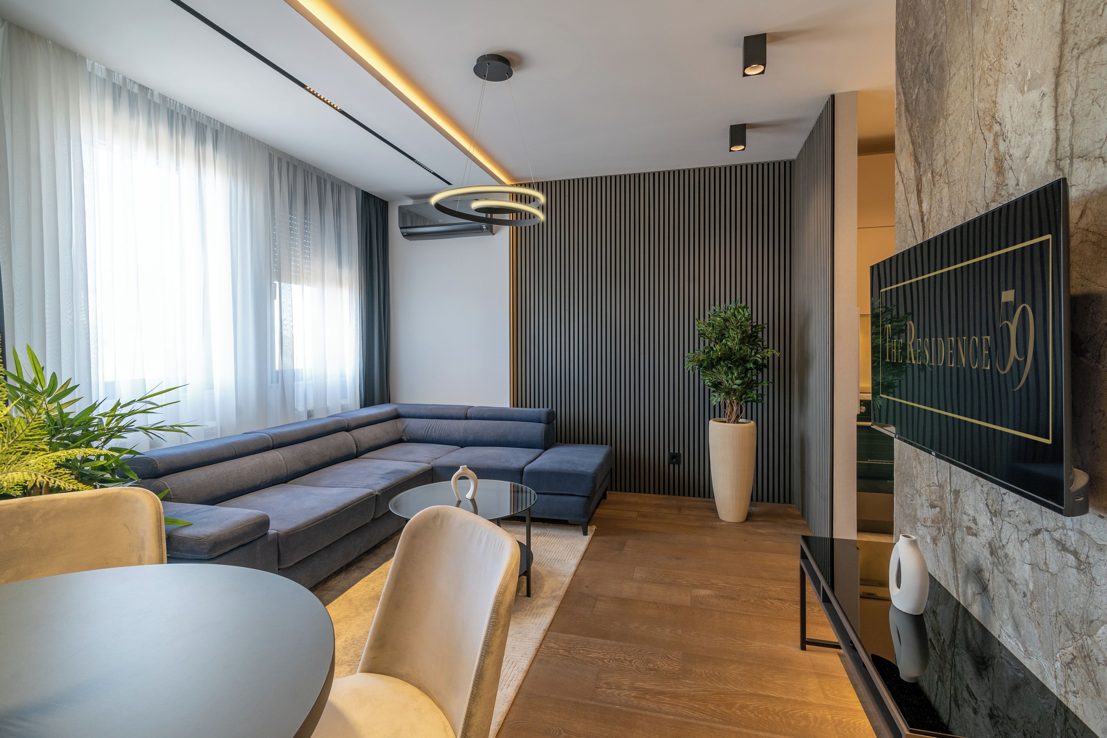 A modern living room with a sectional sofa, a round coffee table, and a dining area with a table and chairs. There is a large potted plant and a wall-mounted TV.