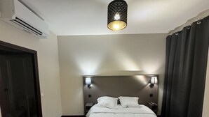 Quarto de hotel com cama, duas luminárias de apoio, iluminação de teto, uma porta e uma janela com cortinas.