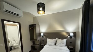 Quarto de hotel com cama, abajures, luminária pendente, espelho e banheiro visível por uma porta aberta.