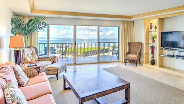 Ein großzügiges Wohnzimmer mit einer großen Couch, einem Couchtisch und einem Fernseher. Ein Balkon bietet Blick aufs Meer.