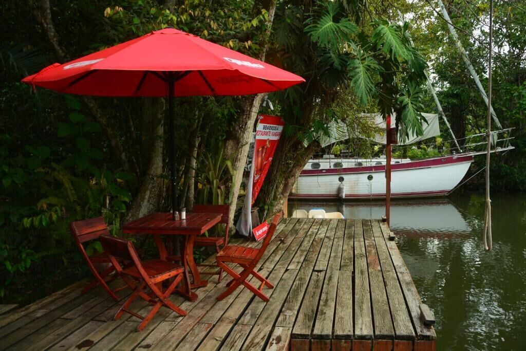 A wooden dock with a red umbrella and a table with chairs, overlooking a body of water with a boat in the background.