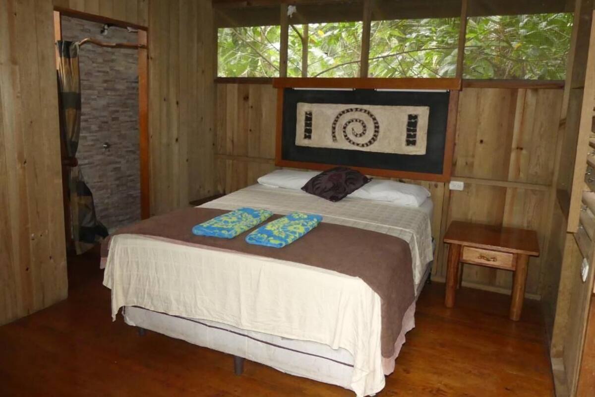 A wooden room with a bed, a small table, and a window with a view of trees.