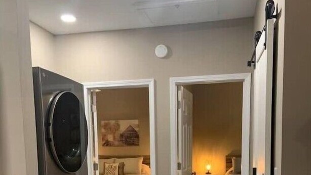 Ein Waschraum mit einer Waschmaschine und einem Trockner, ein Schlafzimmer mit einem Bett und einem Nachttisch sowie ein Flur, der in einen weiteren Raum führt.