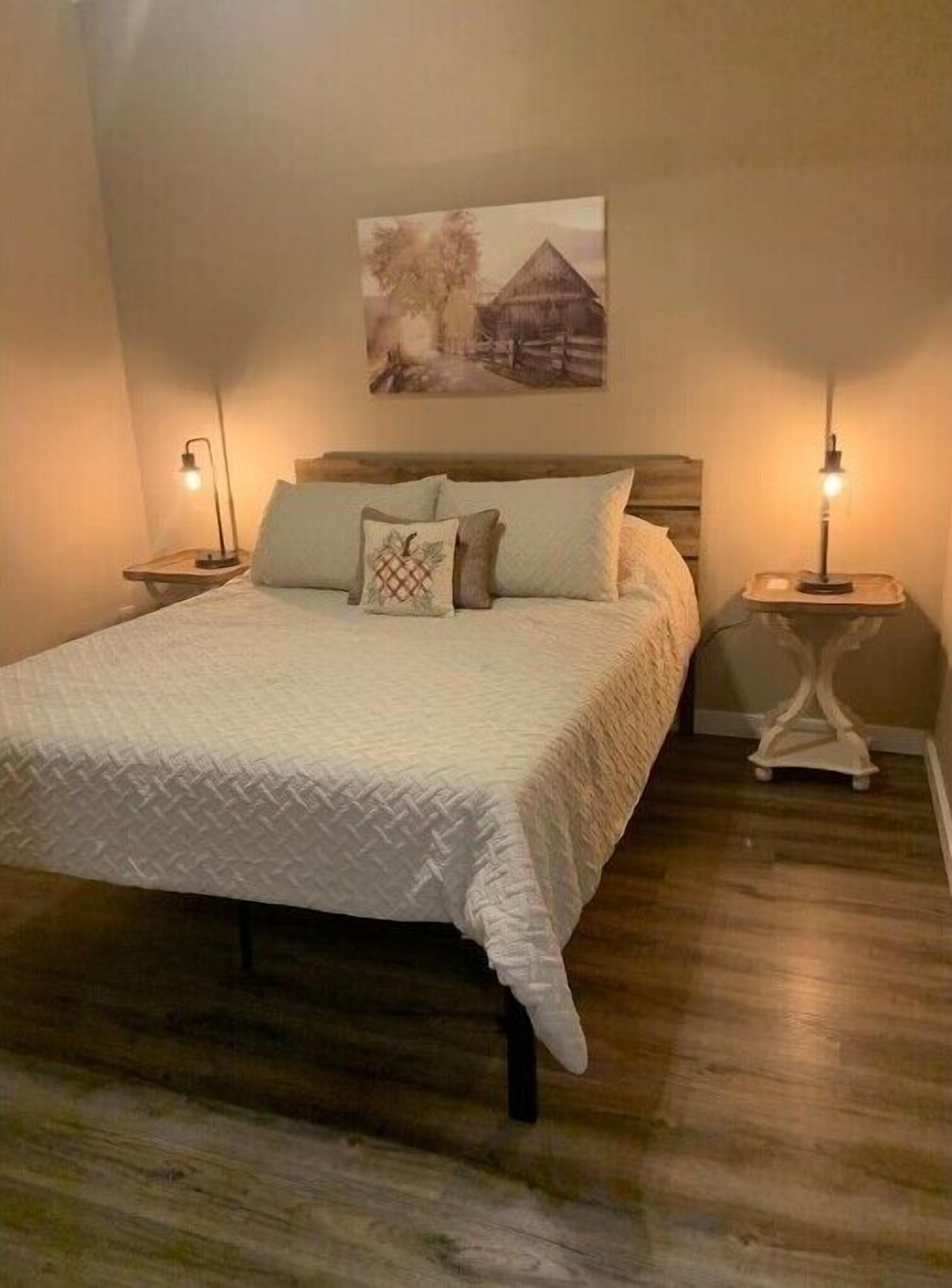 Une chambre à coucher avec un lit en bois, deux tables de chevet avec des lampes, un tableau accroché au mur et un plancher en bois.