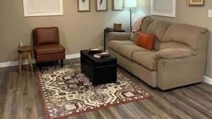 Ein Wohnzimmer mit einem braunen Ledersessel, einem beigefarbenen Sofa mit orangefarbenem Kissen, einem schwarzen Hocker, einem gemusterten Teppich und drei gerahmten Wanddekorationen.