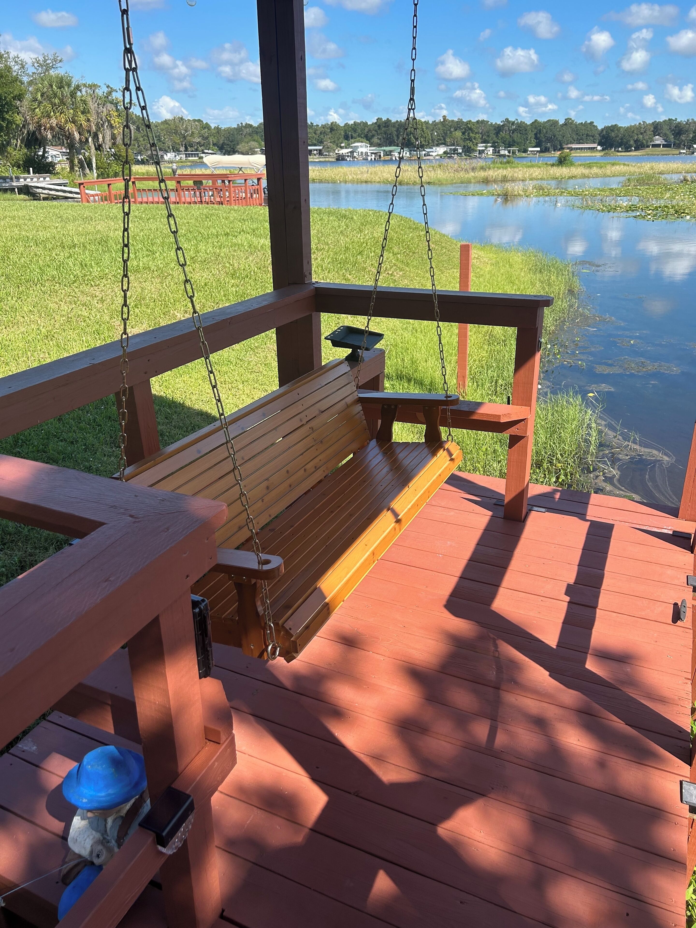 A wooden deck with a bench and a table, overlooking a calm body of water and a grassy area with trees and buildings in the background.
