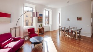 Ein geräumiger Wohnbereich mit einem roten Sofa, einem kleinen runden Tisch, einem Esstisch mit Stühlen und großen Fenstern, die für natürliches Licht sorgen.
