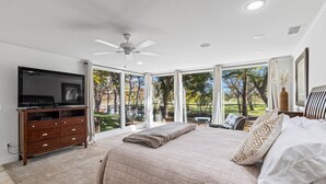 A bedroom with a large bed, a TV, a ceiling fan, and a view of a park. - Turtle Creek House, 4 Bedrooms, 5 Bathrooms, only 8 miles south of Kerrville. (Kerrville)
