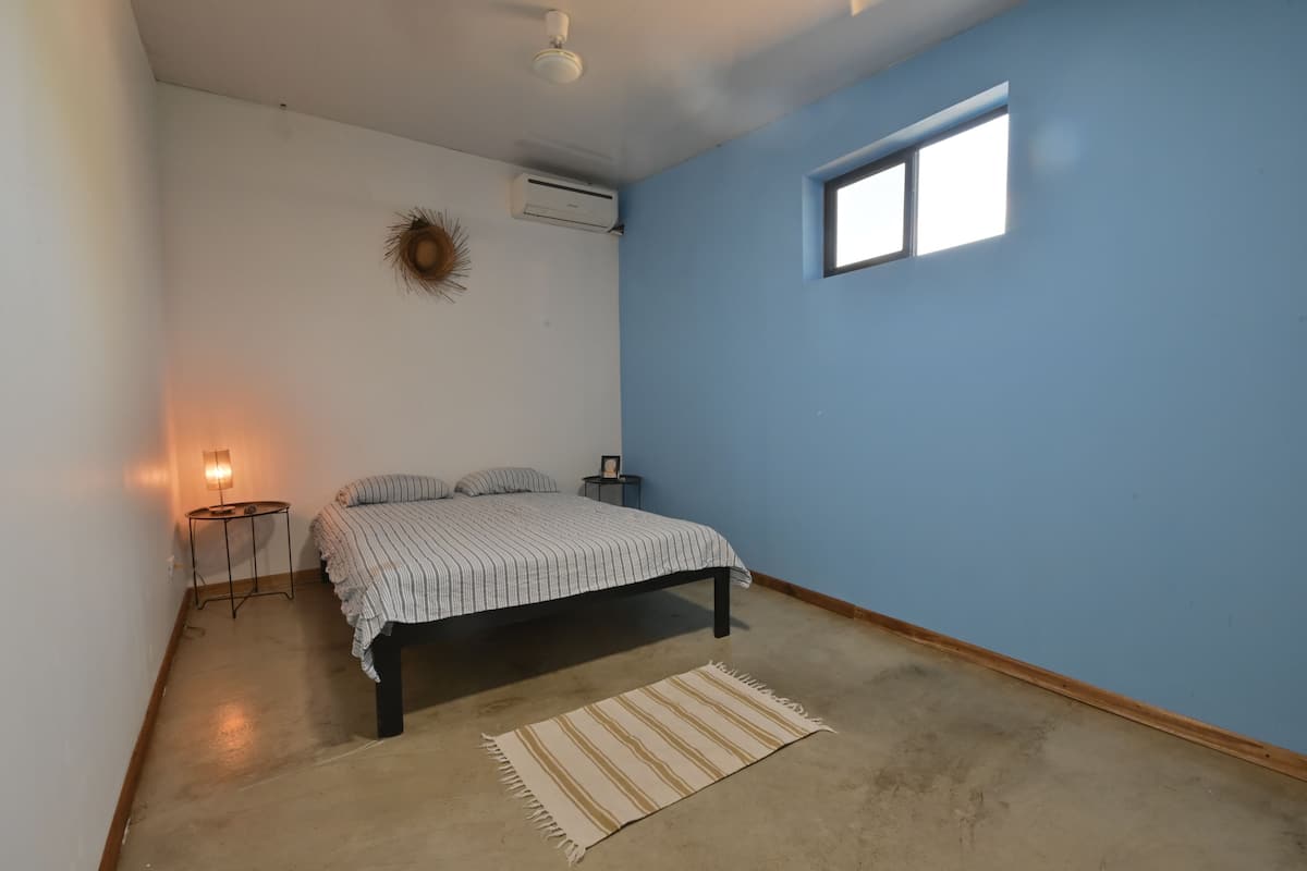 Un dormitorio con una cama, una mesita de noche pequeña, aire acondicionado instalado en la pared, una ventana y una lámpara de pie.