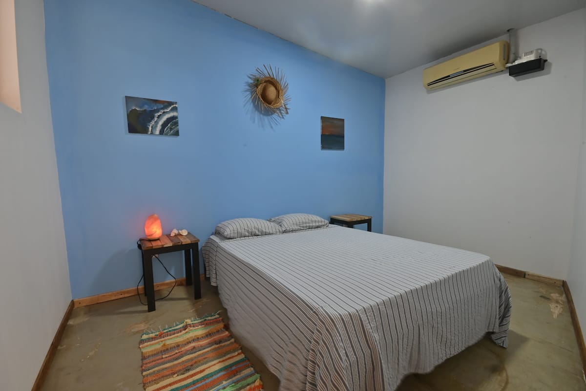 Un dormitorio con cama, mesita de noche, aire acondicionado instalado en la pared y un pequeño adorno en la pared.