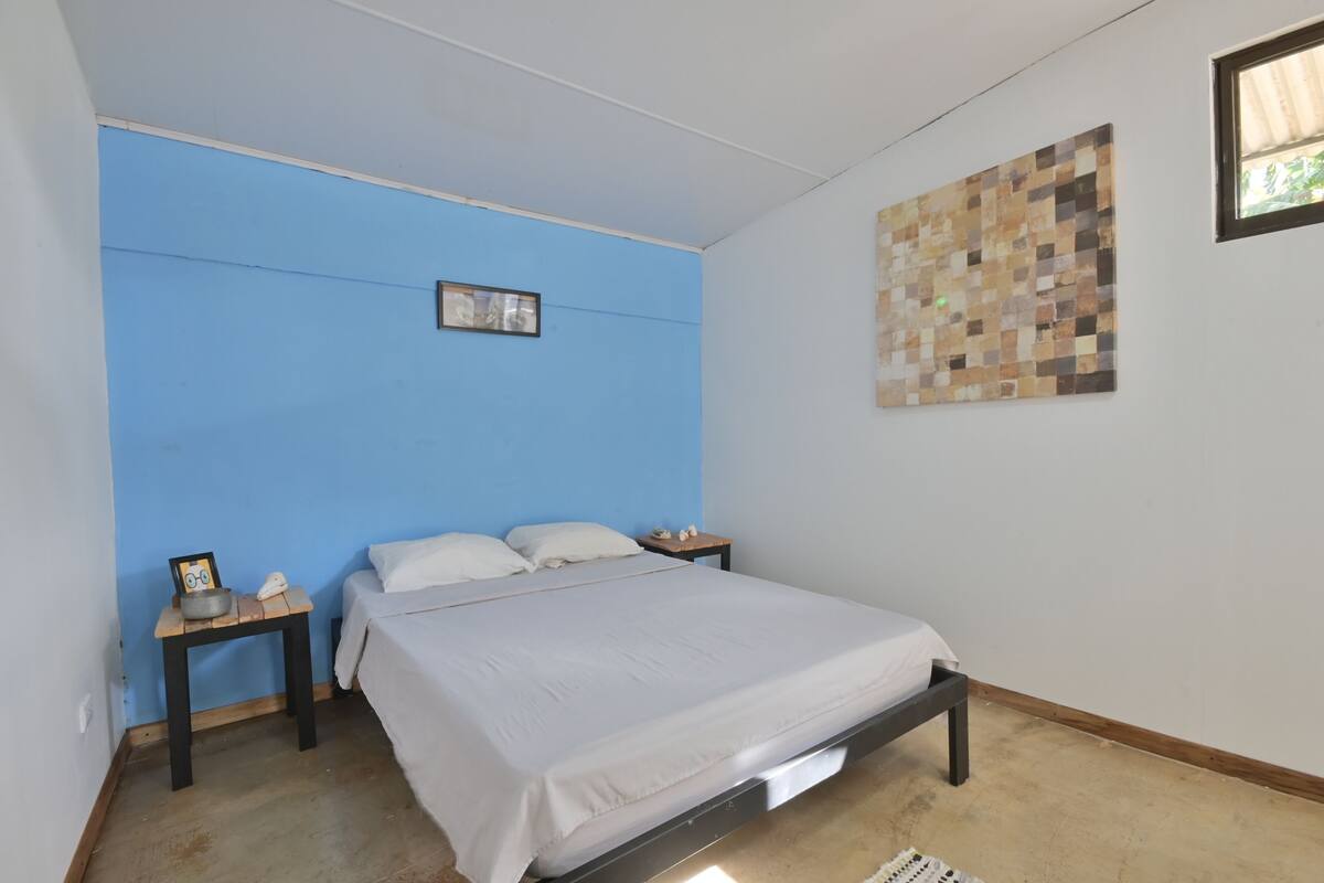Un dormitorio con cama, mesita de noche, reloj de pared y un cuadro enmarco.