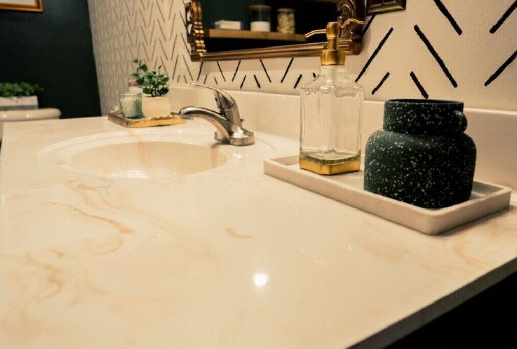 Ein Waschtisch mit modernem Wasserhahn, einer klaren Glasflasche und einem grün strukturierten Behälter auf einer weißen Arbeitsplatte.