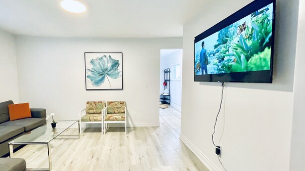 Een moderne woonkamer met een flatscreen televisie aan de wand, een grijze bank, een gestreept fauteuil en een groot bloemrijk kunstwerk.