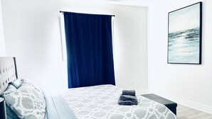 Een slaapkamer met een bed, een bankje, een hoofdeinde, een aan de muur hangend schilderij en een raam met een blauw gordijn.