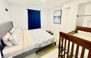 Une chambre à coucher avec un lit, un berceau, un rideau bleu et un tableau encadré au mur.