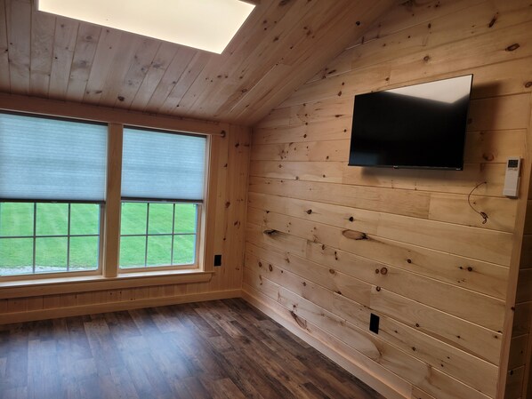 A room with wooden walls, a large window with blinds, a flat-screen TV mounted on the wall, and a wooden floor.