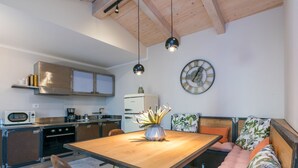 Eine moderne Küche mit Holzausbau, einem Einbauofen, einer Mikrowelle und einem Spülbecken. Ein Esstisch mit Stühlen und eine Bank mit Kissen sind vorhanden. An der Wand hängt eine Zieruhr.