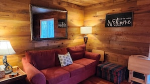 A cozy cabin interior with a wooden ceiling, a red sofa, a plaid ottoman, a small wooden table, and a 'Welcome' sign.