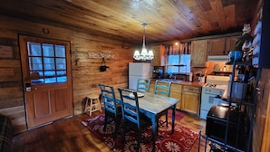 A rustic kitchen with wooden cabinets, a dining table with chairs, a chandelier, and a refrigerator.