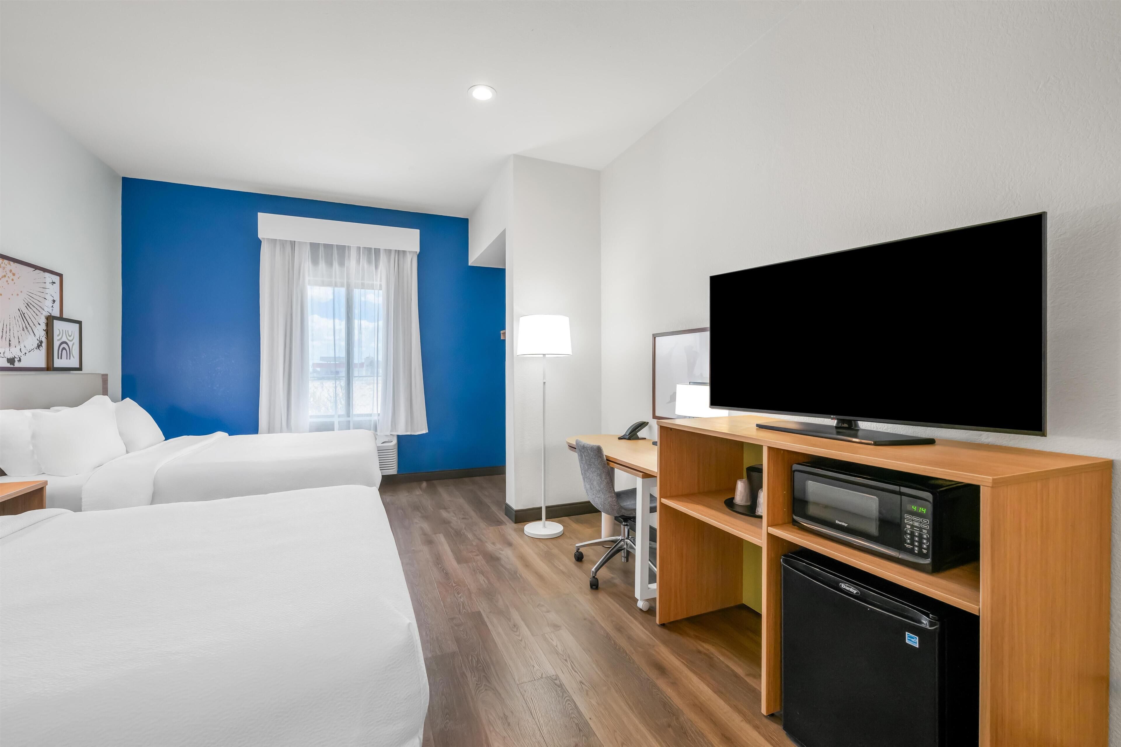 Habitación de hotel con dos camas, un escritorio con computadora, televisor, microondas y refrigerador.