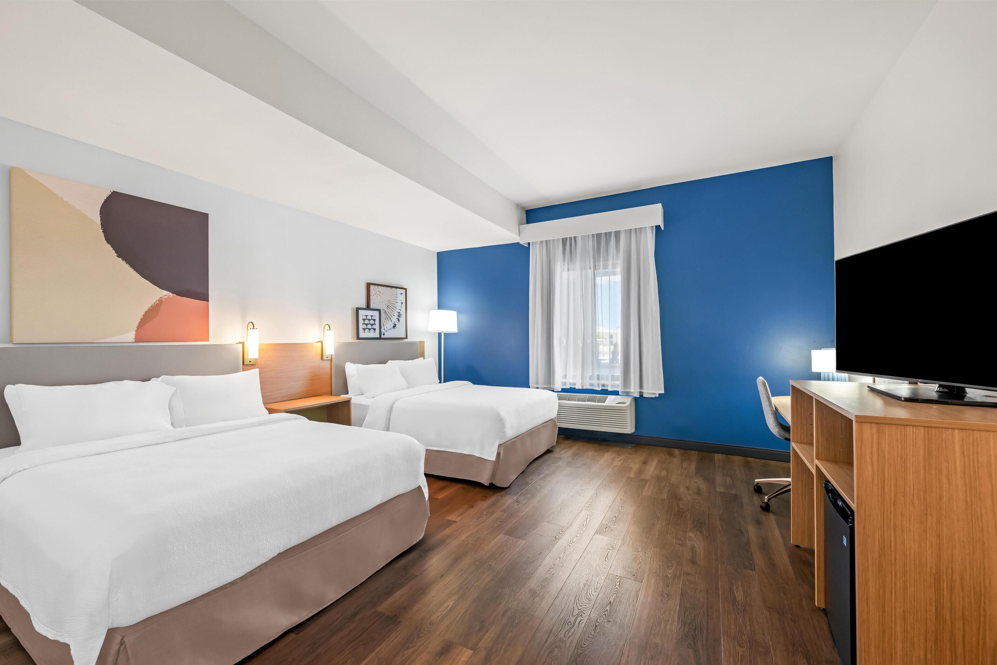 Ett hotellrum med två sängar, ett skrivbord, en tv, ett fönster med gardiner och en tavla på väggen.