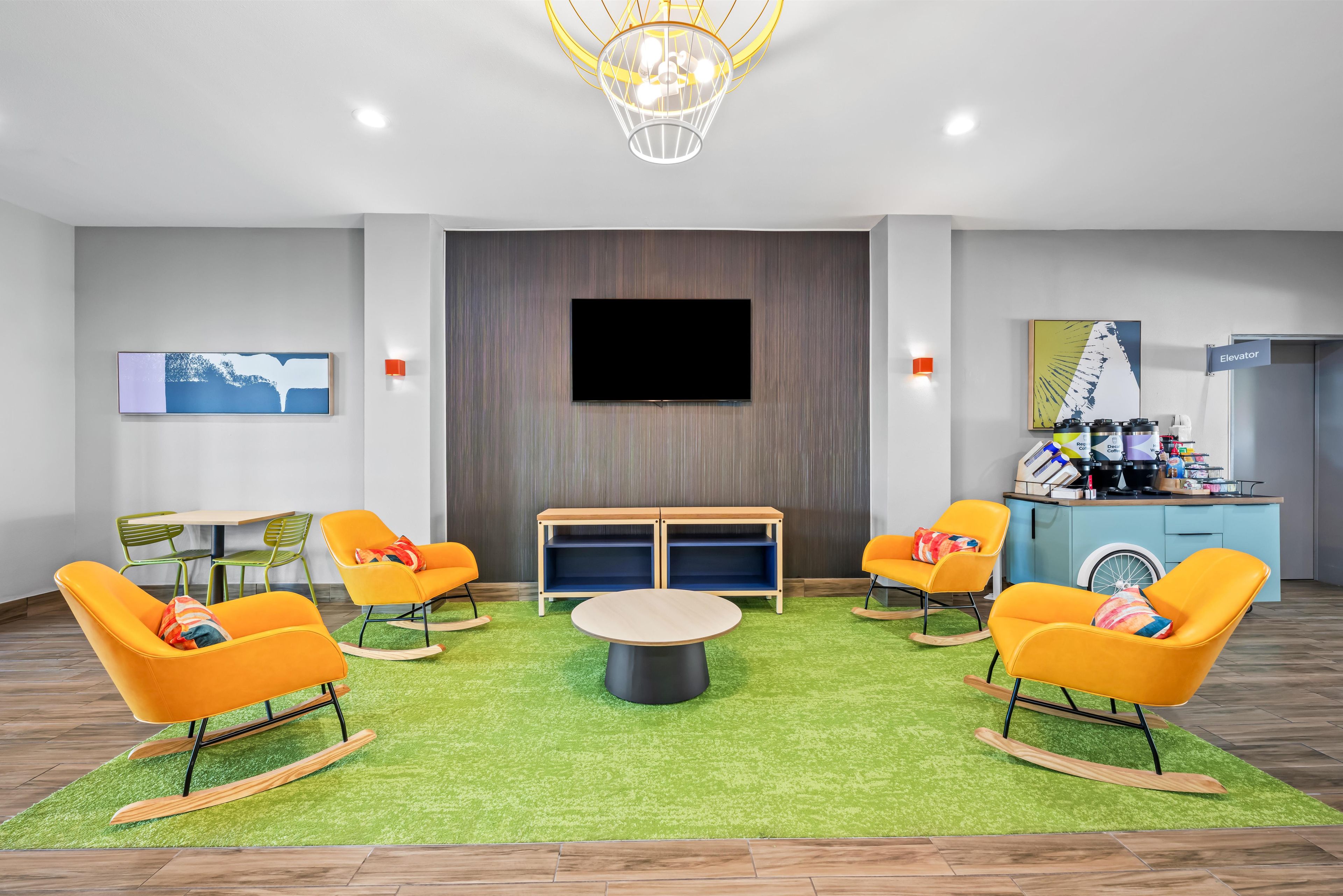 A modern lounge area with a flat-screen TV, rocking chairs, a round coffee table, and a small kitchenette.