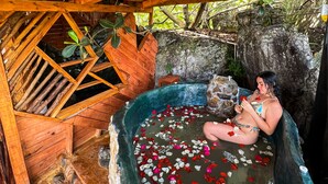 Une personne se détend dans une baignoire en pierre remplie d’eau et de pétales de rose, sous une structure en bois surmontée d’un toit de chaume.