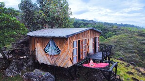 Une cabane rustique en bois, dotée d’un toit de chaume, d’un balcon orné d’un tissu rouge et offrant une vue sur une colline verdoyante et boisée.