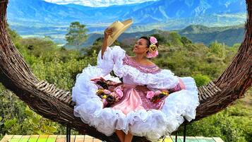 A person in a traditional dress sitting on a colorful wooden bench with a heart-shaped woven structure behind them, overlooking a mountainous landscape.