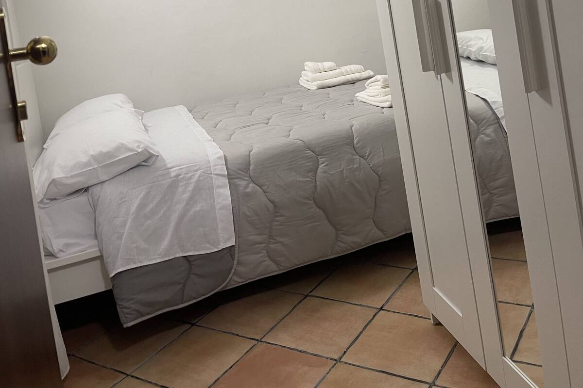 A small, neatly made bed with white linens and a grey quilt, a closed wardrobe, and a tiled floor.