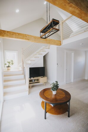 Um amplo loft com teto alto, vigas de madeira aparentes, escada, televisão de tela plana e uma mesa de centro redonda de madeira com uma planta.
