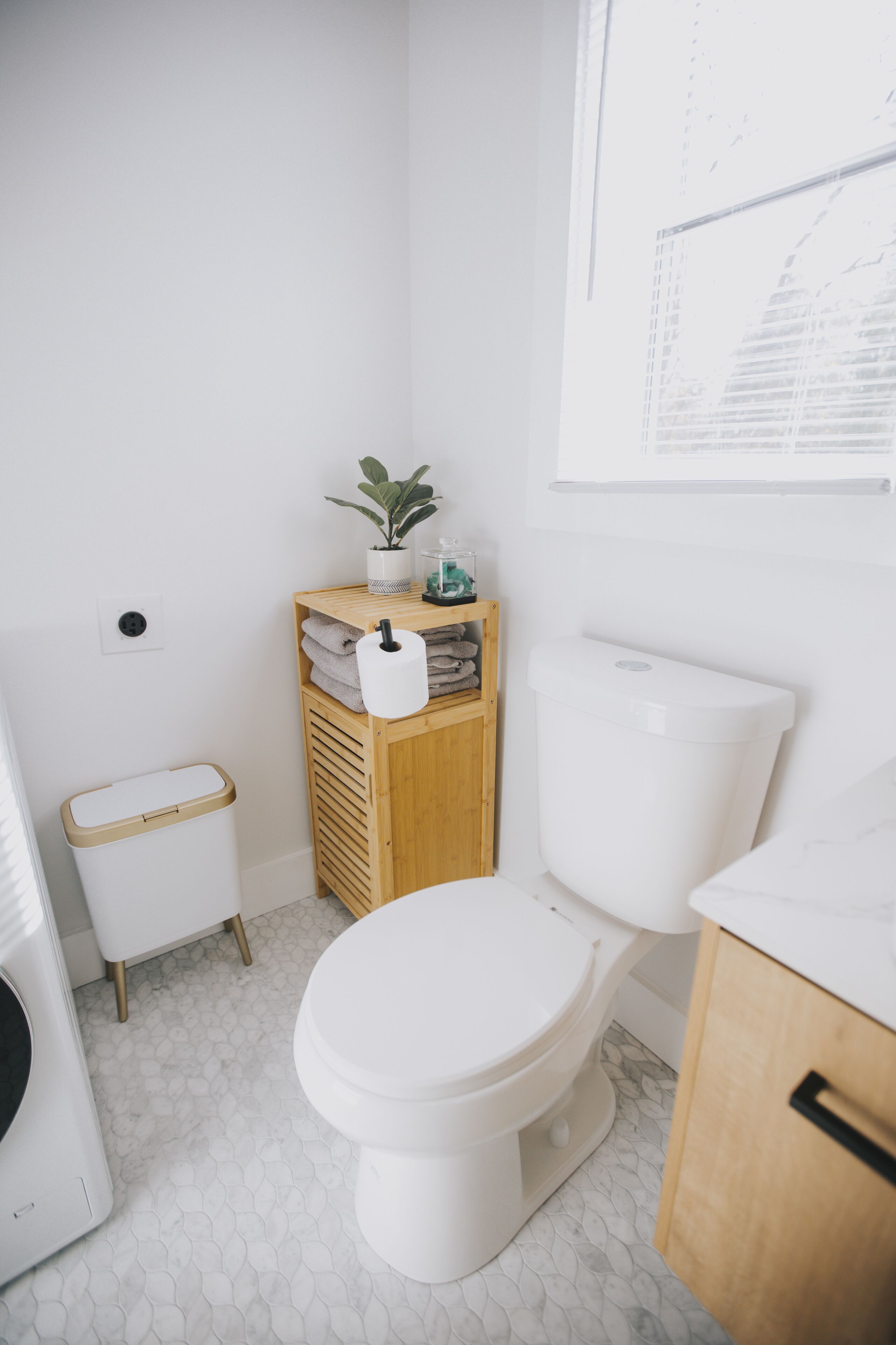 Une petite salle de bain moderne comprenant des toilettes, une étagère en bois avec des serviettes, une poubelle et une petite plante.